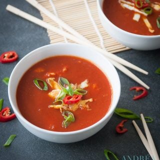 A grey background with two bowls filled with Asian tomatosoup with egg, spring onions and pepper. Some chopsticks and veggies scattered around