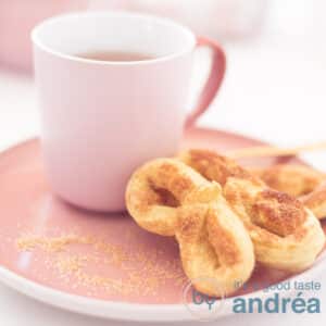 A square photo with a pink plate with three pretzels and a pink cup filled with tea.