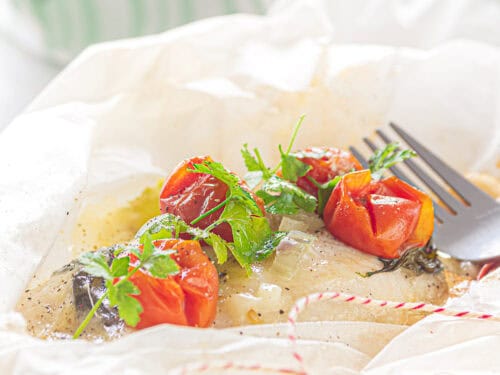 A square photo with an open baking paper filled with fried cod with cheese and vegetables
