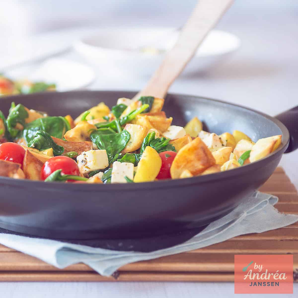 A square photo showing part of a wok filled with stir-fried potatoes, feta, spinach and tomato.