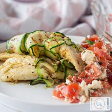 A square photo with a white plate on which is a tilapia fillet rolled in zucchini with a delicious tomato salad.