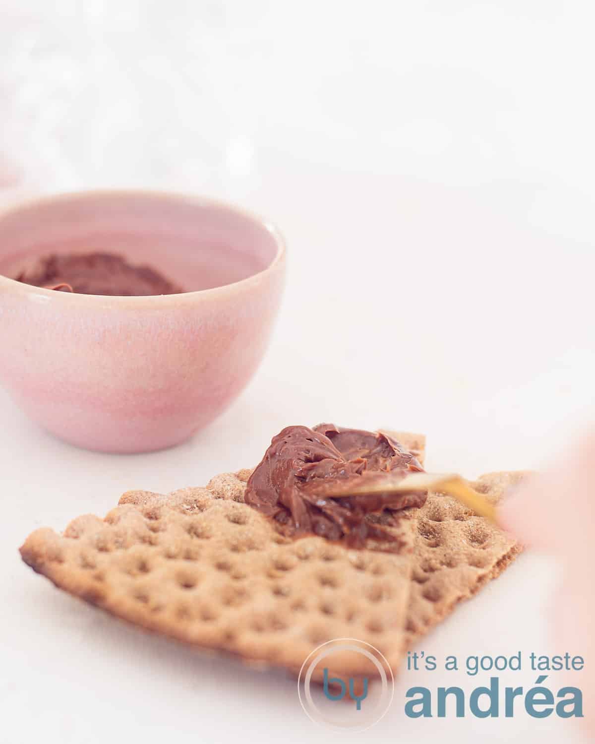A person is smearing with a knife chocolate butter on toast. A pink bowl in the back.