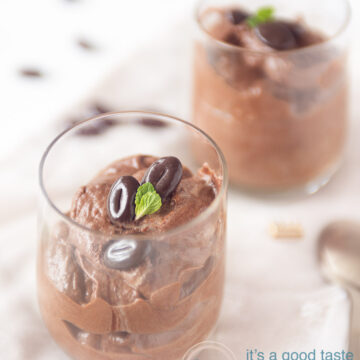 Two glasses filled with a mousse of dark chocolate. Garnished with chocolate beans and mint leaves. Some chocolate scattered around