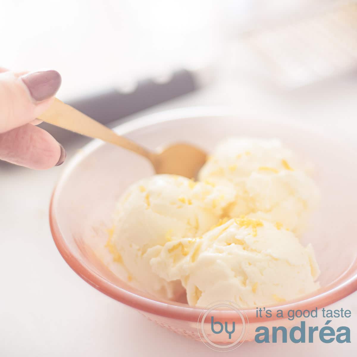 A square photo with homemade lemon ice cream in an orange bowl. A spoon takes a bite. A grater in the background