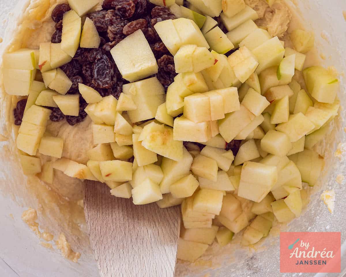 Een glazen kom met muffinbeslag waaraan appelblokjes en rozijnen zijn toegevoegd.