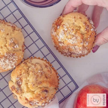 A square picture of three apple muffins with raisins. A hand takes one.