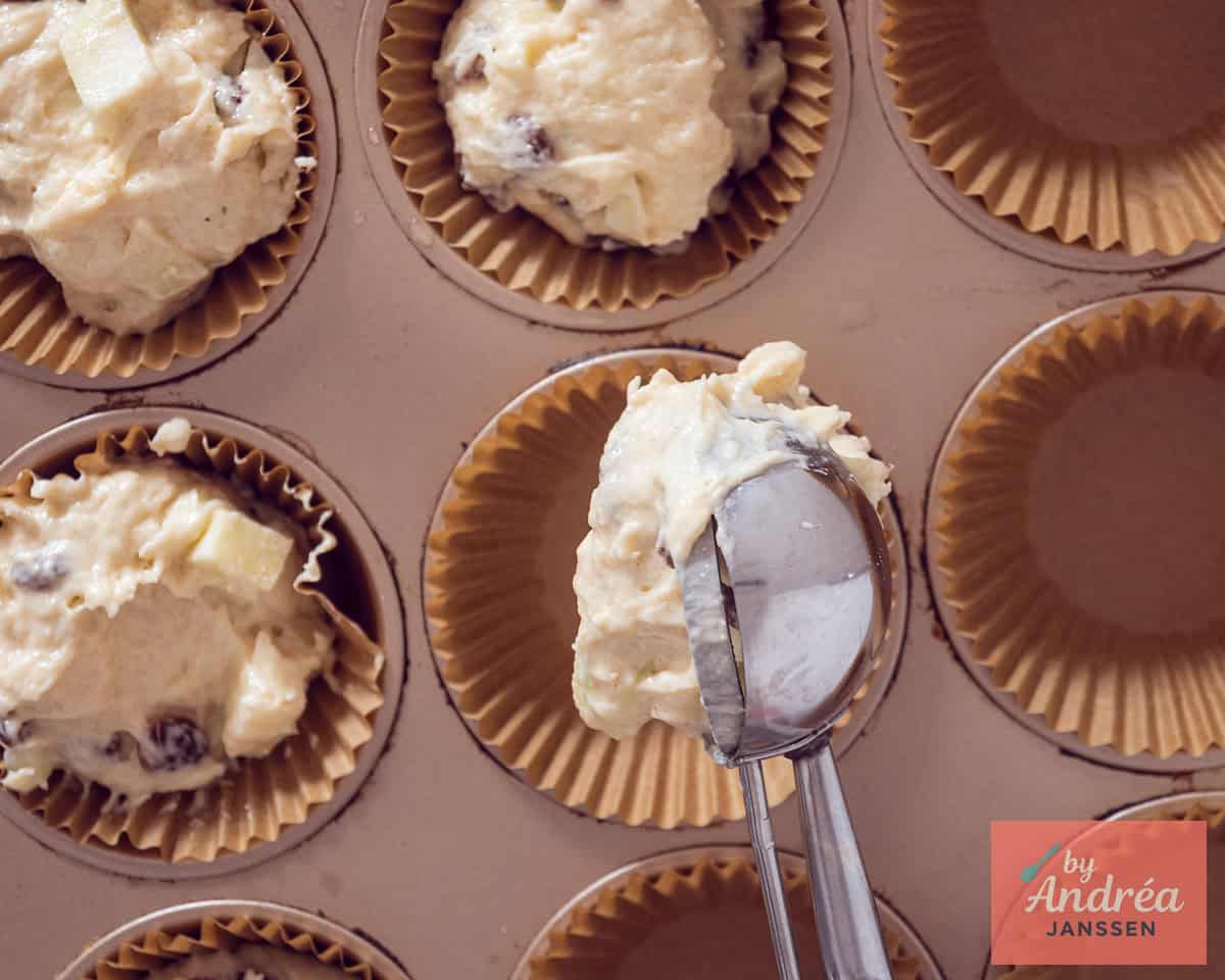 Een muffinvorm waarin cupcakevormpjes appel muffin met rozijnen beslag wordt gedaan.