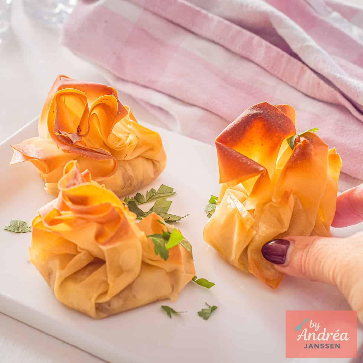 A square picture with a white plate with three Goat Cheese Phyllo Appetizers. A hand taking one.
