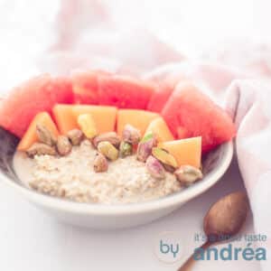 A square photo with a gray bowl with oatmeal, pistachio, two kinds of melon on a white background. A pink white tea towel in the background
