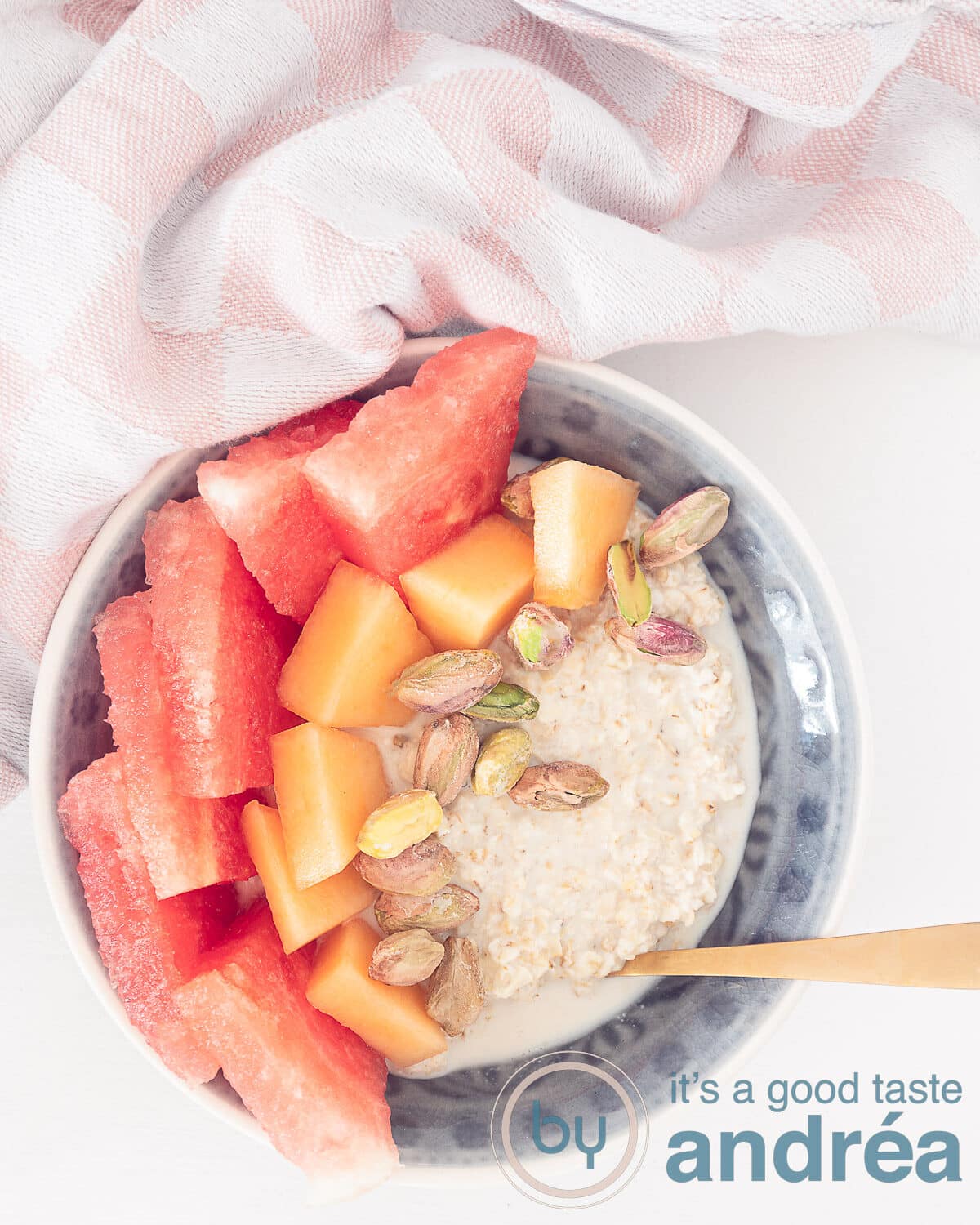 Een foto van bovenaf met een grijs schaaltje gevuld met overnight oats met twee soorten meloen en pistache op een witte ondergrond. Bovenin ene roze witte doek.