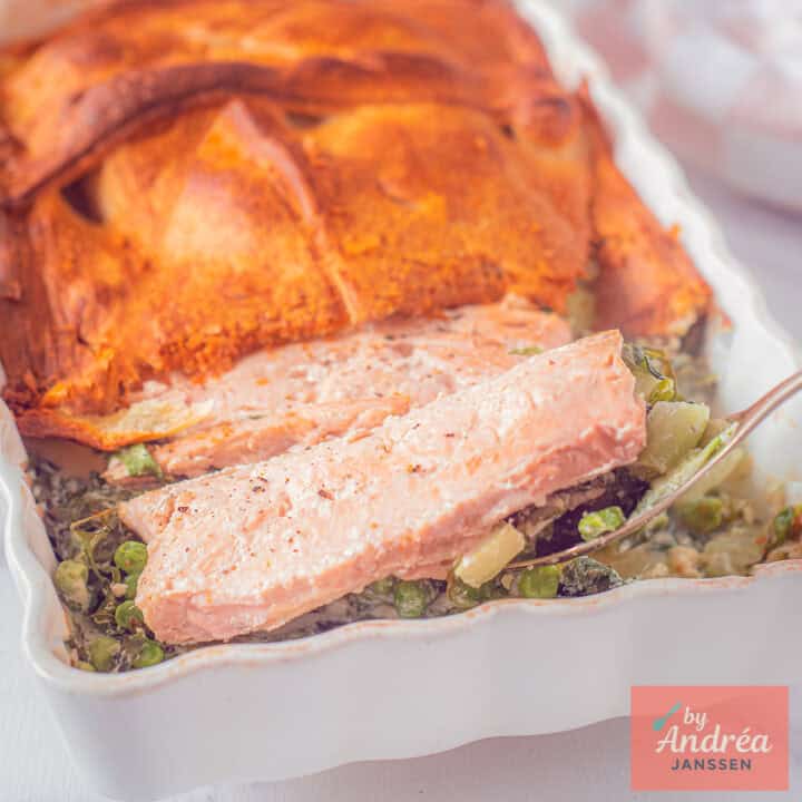 Een vierkante foto met een ovenschotel met groene groenten, zalm en een laagje croissantdeeg.