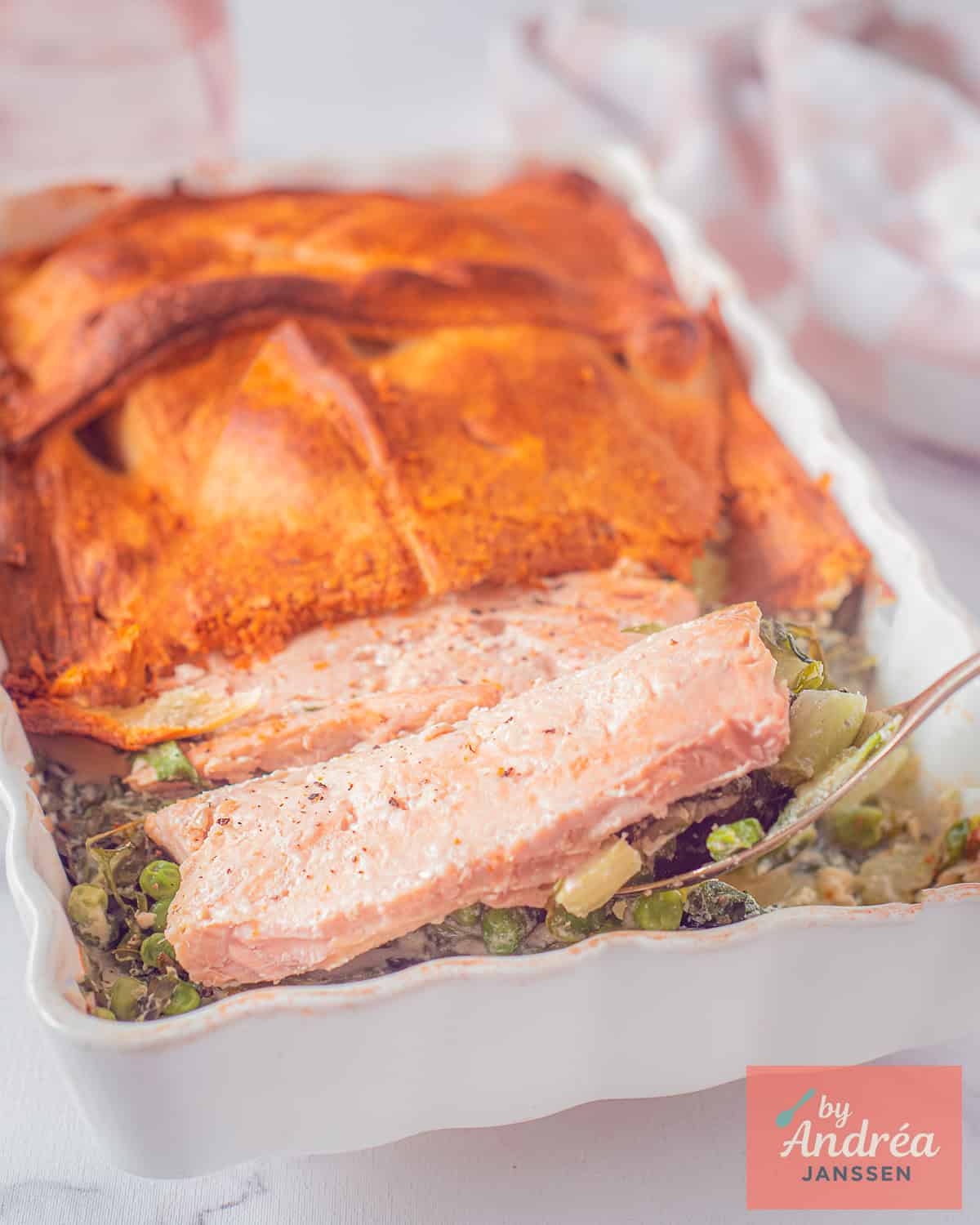 Een witte ovenschotel met stukken zalm, croissantdeeg en groenten. Een lepel schept de zalm omhoog.