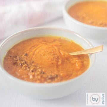 A square photo of a blue bowl filled with carrot curry soup. A golden spoon lies in the soup.