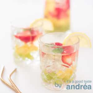 A square photo with two glasses filled with white sangria with fruit on a white background. On the left part of two golden straws.