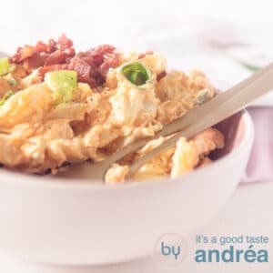 A square photo with a pink bowl filled with kartoffel salad, bacon and green onion on a white background