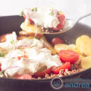 A square shot of a black skillet filled with a Greek stew with minced meat and yogurt cheese topping. A spoon takes out a bite.