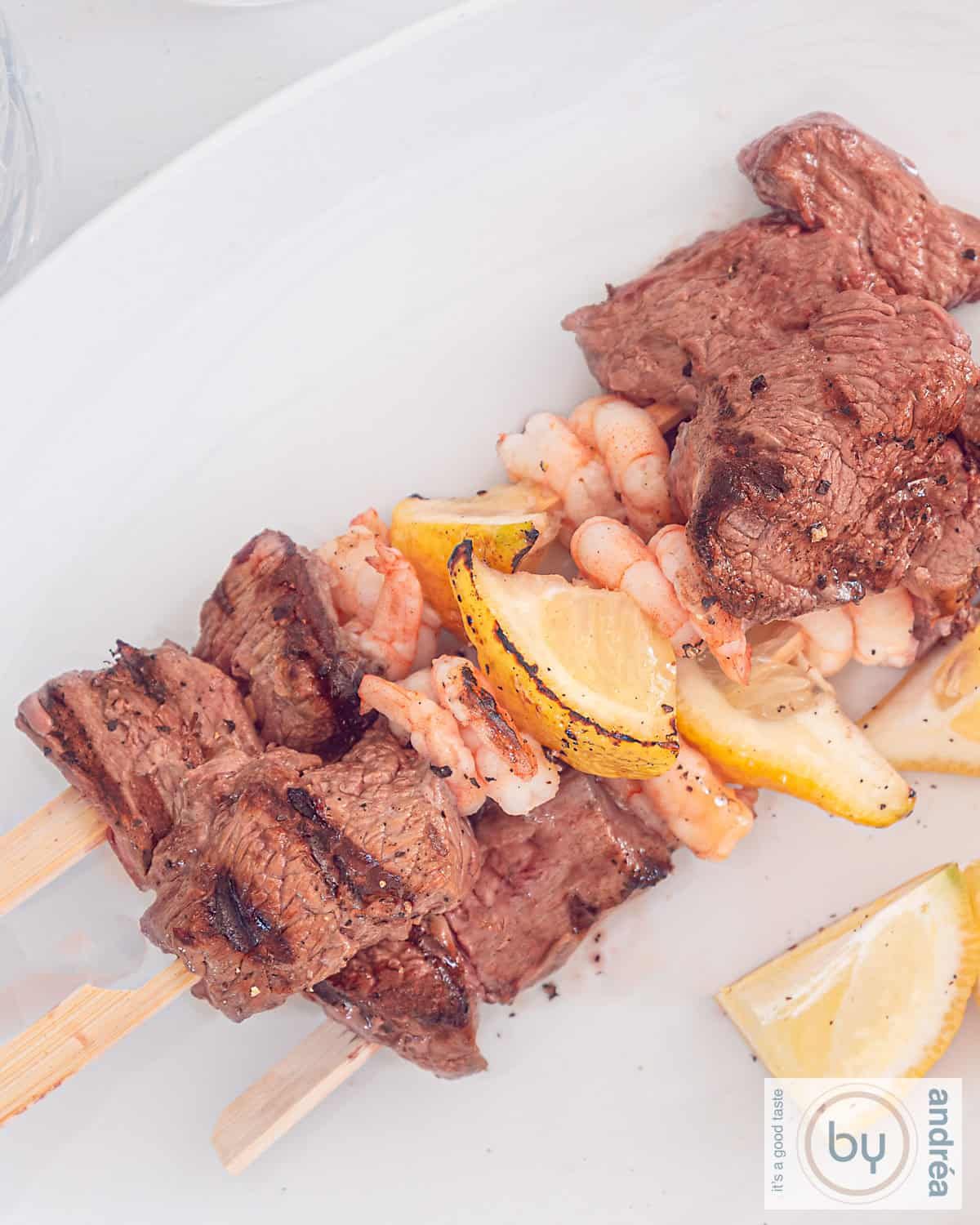 A photo from above: three skewers on a white plate with steak, shrimp and lemon