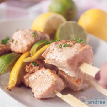 A square photo with a white bowl with three skewers of salmon, lime and lemon. A hand picks up a skewer