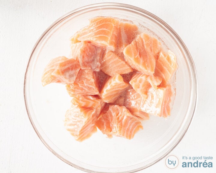 A glass bowl with a lemon, lime, gin marinade in which cubes of salmon have been mixed.