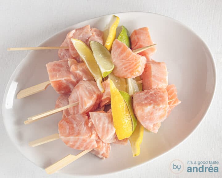 A white plate with five skewers with cubes of salmon, lemon and lime wedges strung on a skewer.