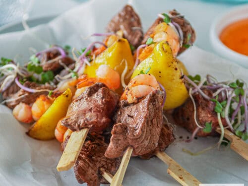 A square photo with a white background with five skewers of surf and turf steak with shrimp. A bowl of chili sauce and a blue cloth in the background.