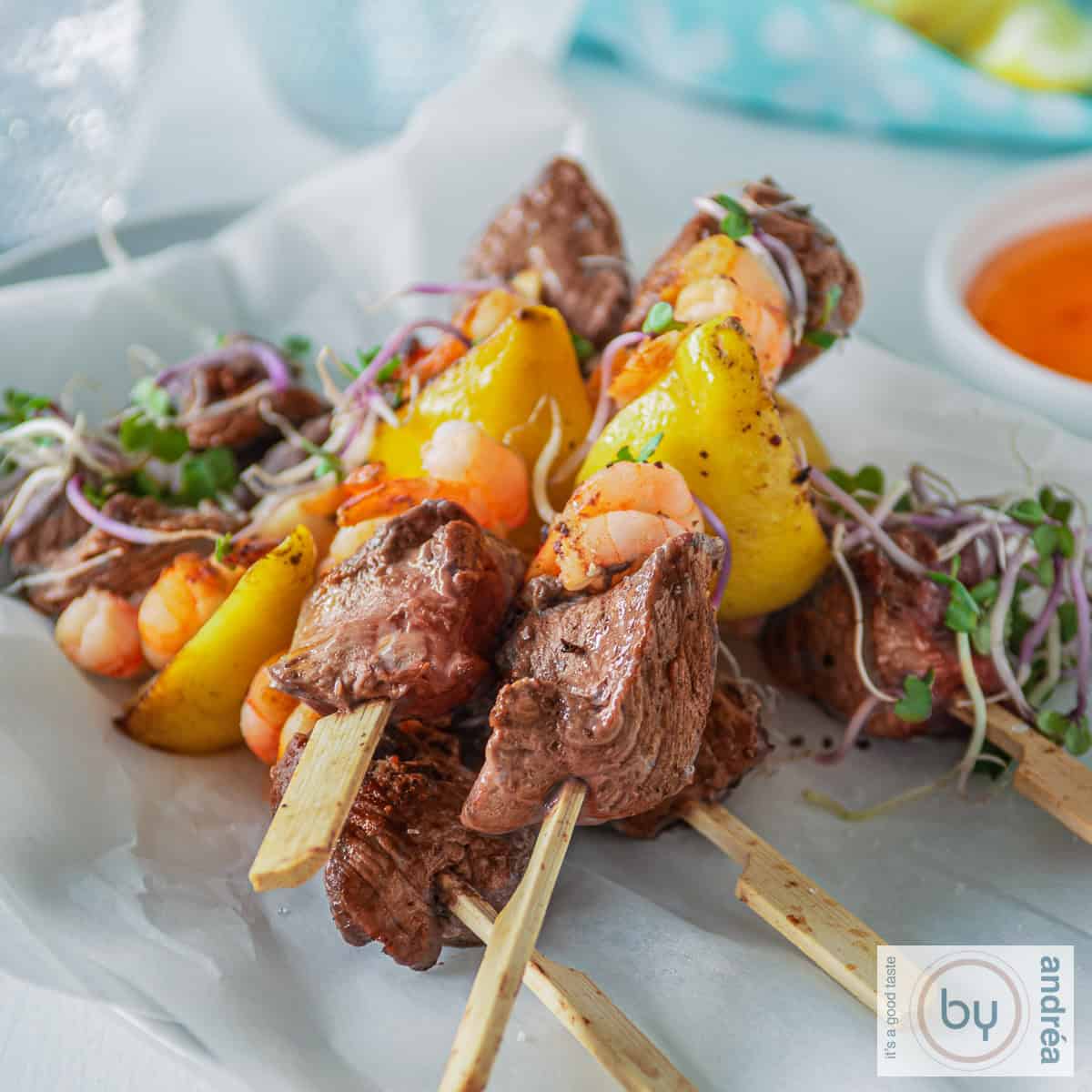 A square photo with a white background with five skewers of surf and turf steak with shrimp. A bowl of chili sauce and a blue cloth in the background.