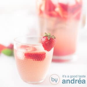 A square photo with a glass of strawberry lemonade with fresh strawberries in the front and a can of lemonade in the back