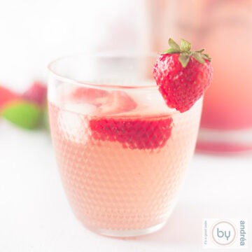 A square photo with a glass of strawberry lemonade with ice and fresh strawberries in the foreground.