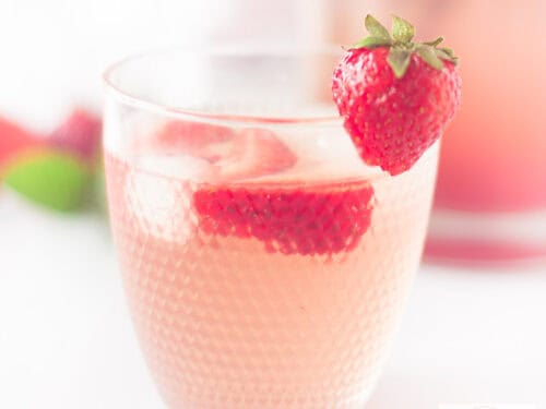 A square photo with a glass of strawberry lemonade with ice and fresh strawberries in the foreground.