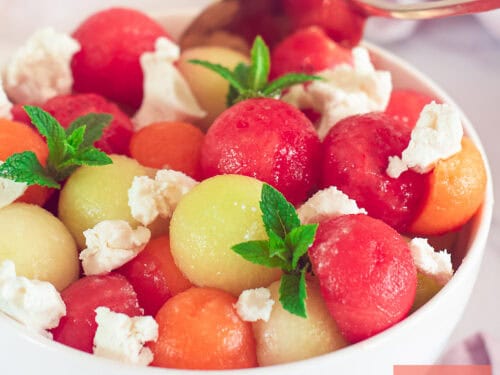 A square photo of a white bowl of galia, cantaloupe, and watermelon balls with goat cheese and mint.