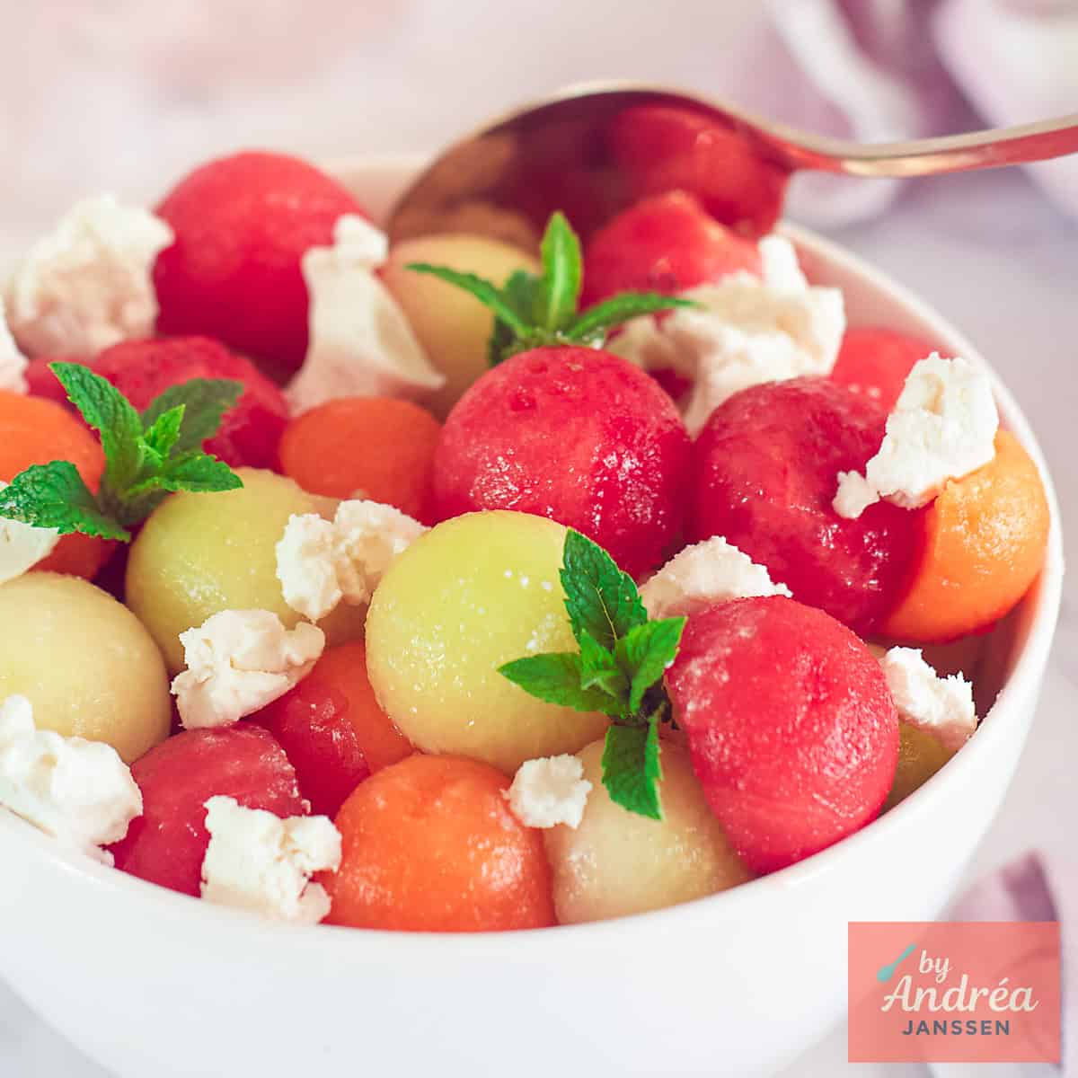 A square photo of a white bowl of galia, cantaloupe, and watermelon balls with goat cheese and mint.