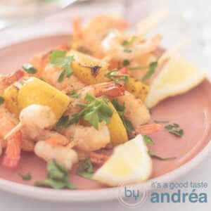 A square photo with a pink plate with three shrimp skewers interspersed with lemon and garnished with parsley.