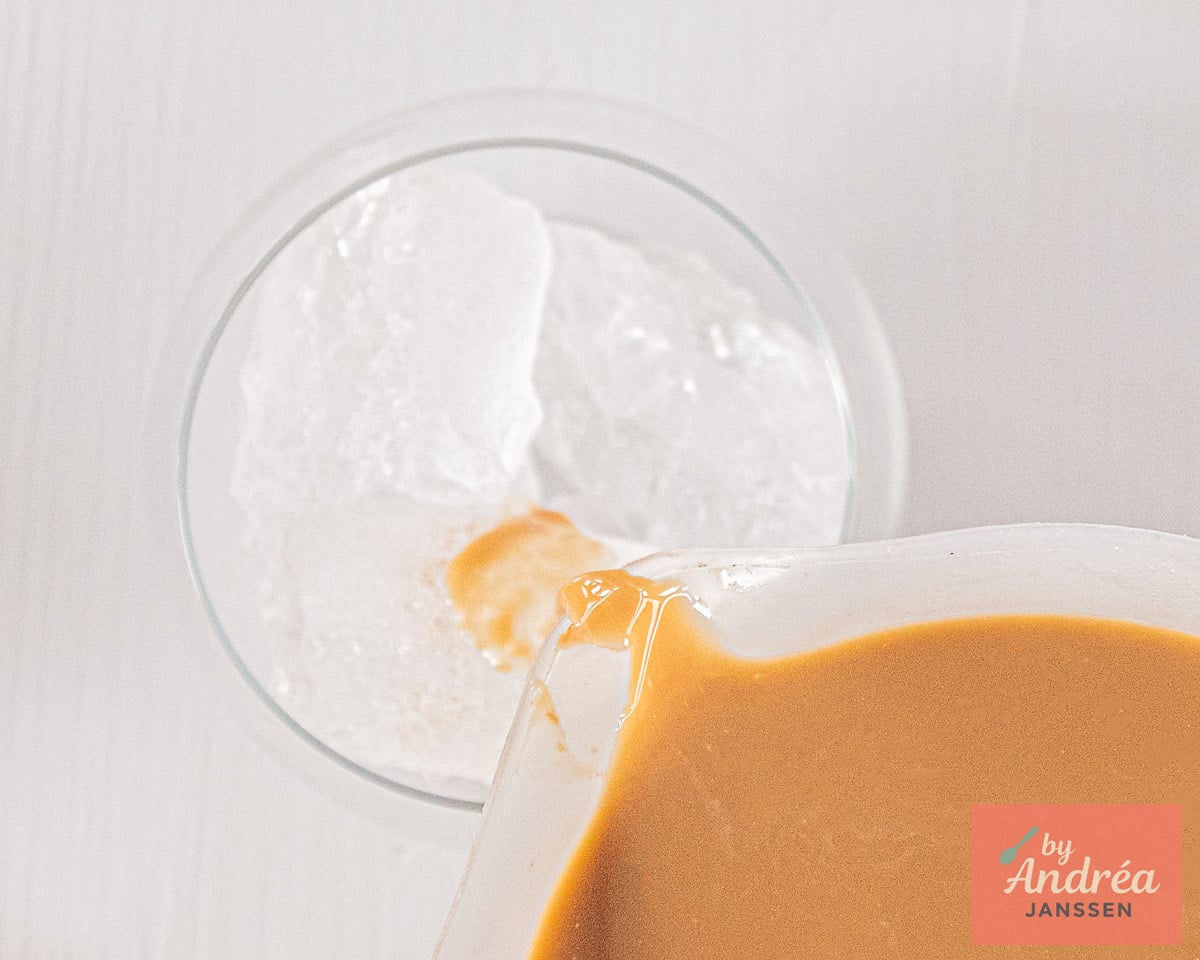 The creamy coffee mixture being poured over ice cubes in a glass