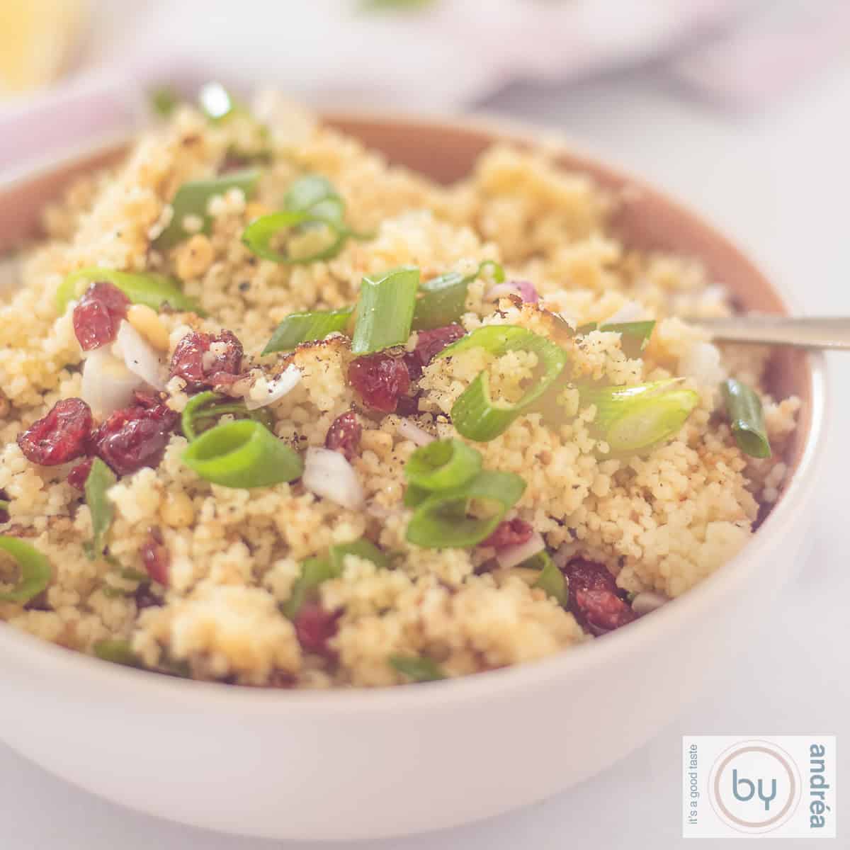 A square photo of a bowl of couscous with pine nuts, lemon and cranberries