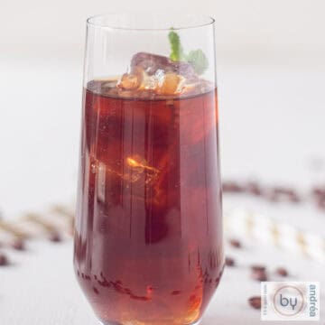 A square photo of a glass filled with Mexican iced coffee on a white surface. Coffee bean and straws lie around it
