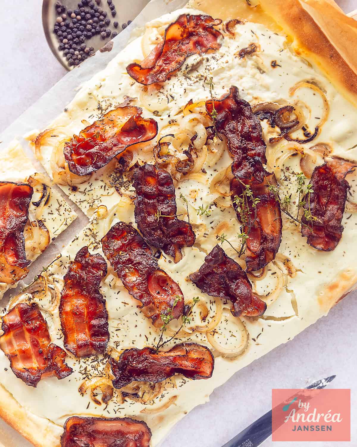 A top-down photo of a tart with flammkuchen, fried bacon, onion, thyme, and crème fraîche