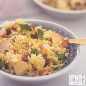 A square photo with a gray bowl filled with a rice salad with curry, smoked chicken, raisins and walnuts