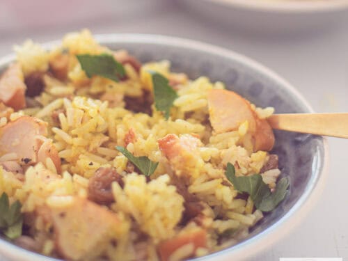 A square photo with a gray bowl filled with a rice salad with curry, smoked chicken, raisins and walnuts
