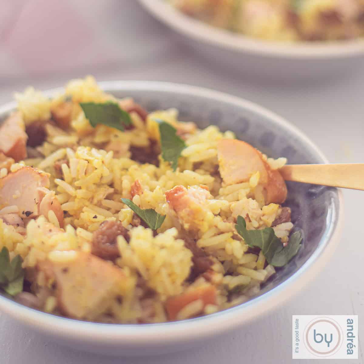 A square photo with a gray bowl filled with a rice salad with curry, smoked chicken, raisins and walnuts