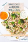 A white plate on a white background with rice baked with broccoli, egg and peas. At the bottom two chopsticks, on the right side a bowl with chili sauce. At the top a text overlay: dinner, broccoli and egg fried rice, easy recipe, vegetarian.