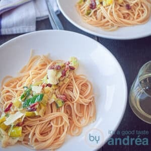 A square photo with a white plate with spaghetti, mascarpone, bacon and fried leeks on it on a dark background. In the background a second sign.