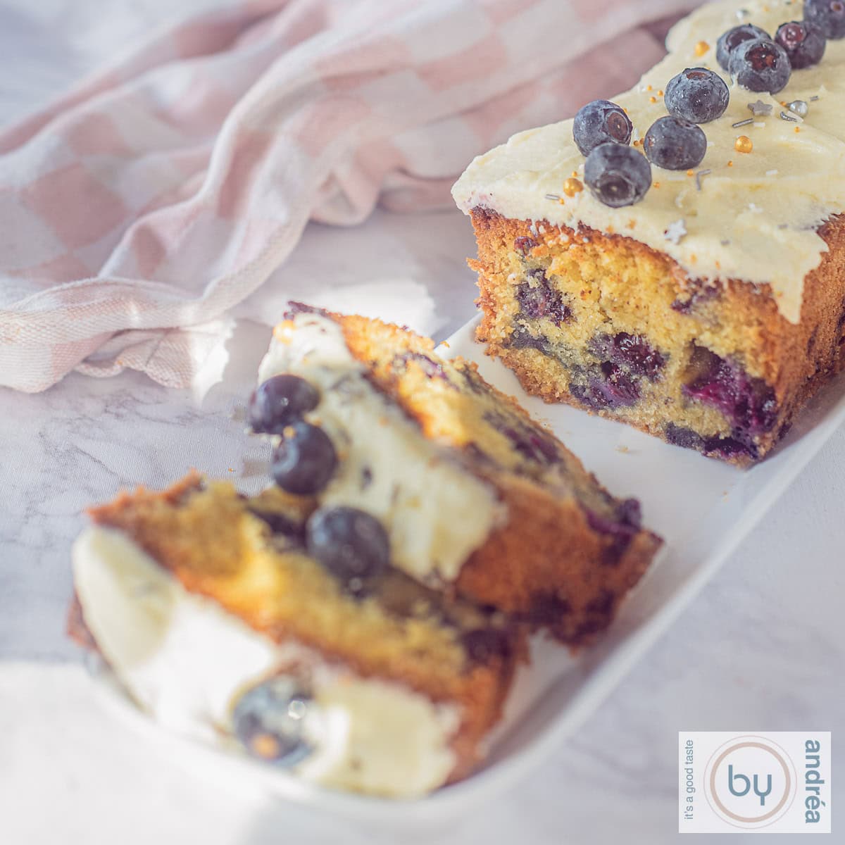 A square photo of a slice of blueberry cake with vanilla frosting. Two slices have been cut off.