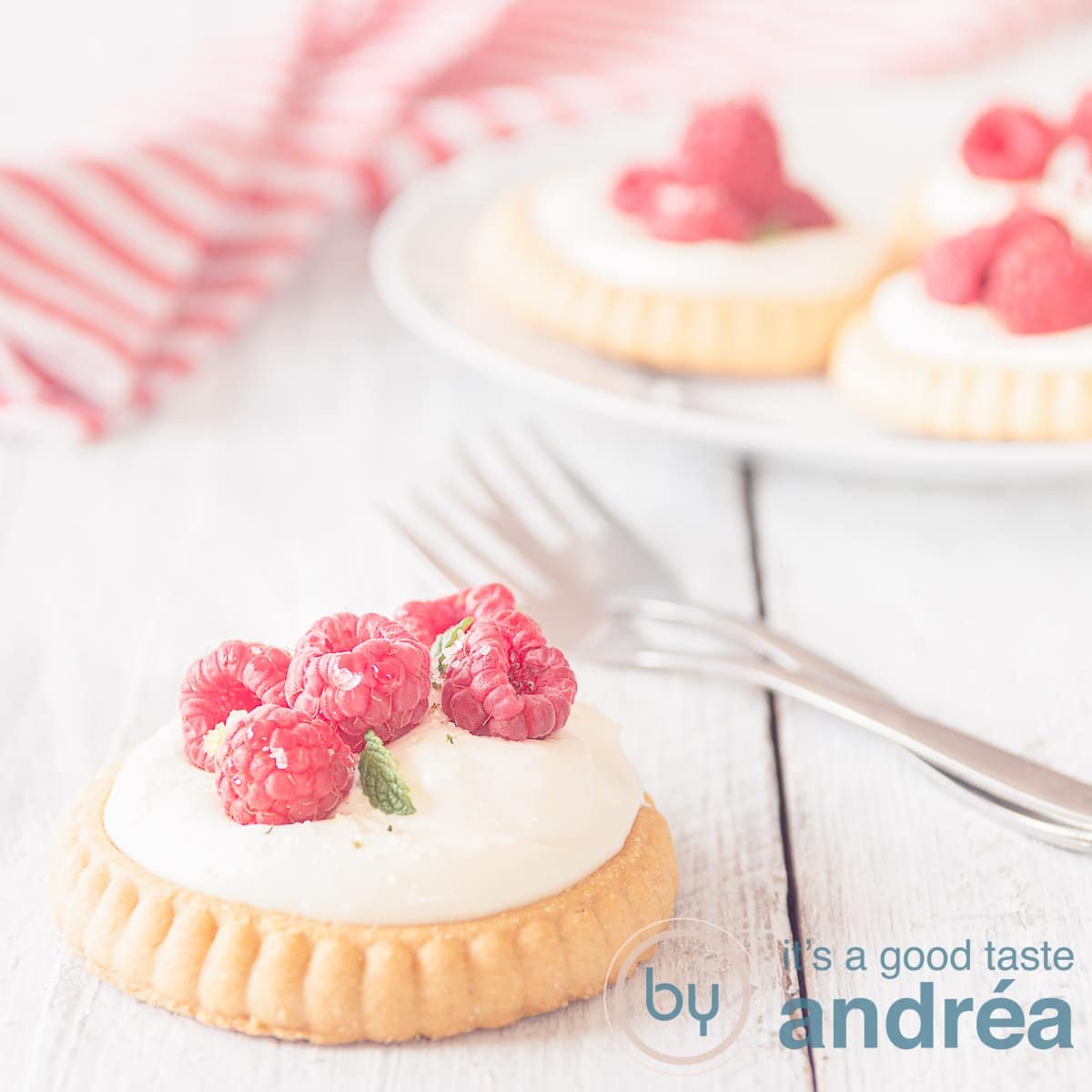 A square photo of four raspberry pastries, one in the foreground on a white wooden background. Two forks are ready. A white red striped tea towel in the background