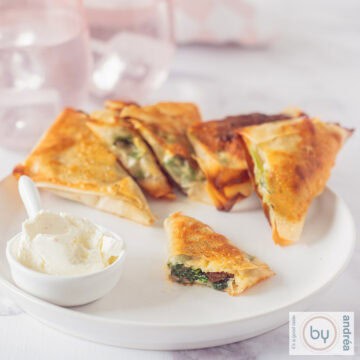 A square photo of a white plate with filo pastry spinach snacks in a row. A dip is diagonally in front of it. One of the snacks is halved so you can see the filling.