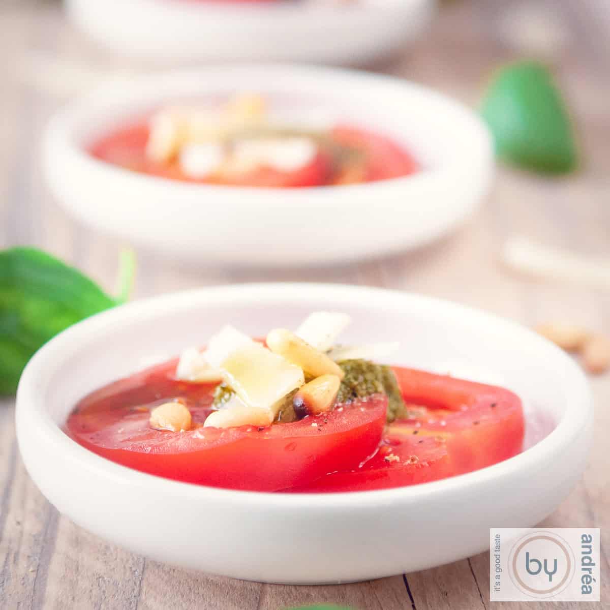 A square photo with a wooden background with three white bowls filled with tomato slices, grated cheese, pesto and pine nuts. Cheese and basil are around it.