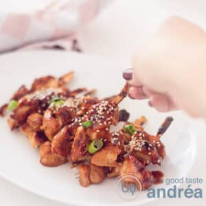 A square photo with a white plate filled with yakitori chicken skewers garnished with spring onion and sesame seeds. A hand takes a skewer