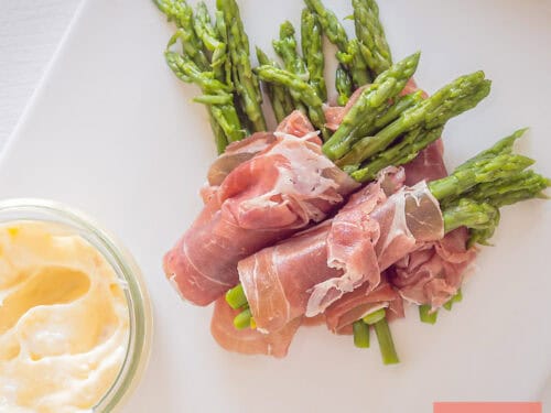 A square picture taken from above with a white plate with homemade aioli and green asparagus in parma ham appetizers