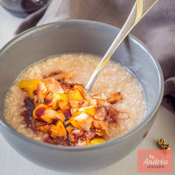 A square photo with a gray bowl of oatmeal, mango, date syrup, and coconut