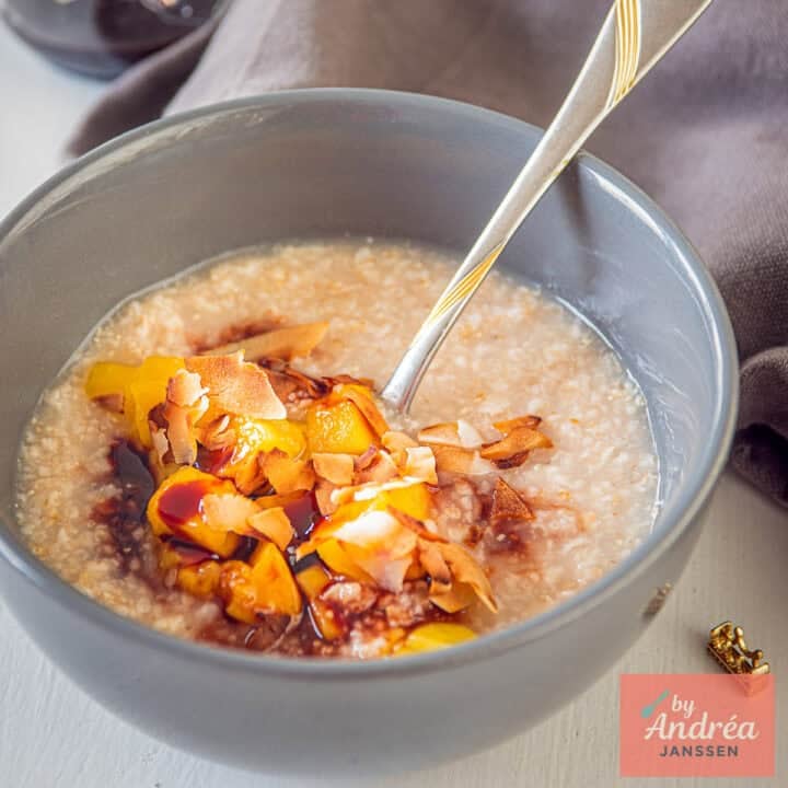 A square photo with a gray bowl of oatmeal, mango, date syrup, and coconut