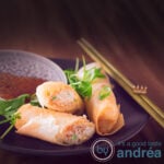 A square photo with two spring rolls on a black plate. A third chicken spring roll dipped in a sauce. In the background a wicker roll mat.